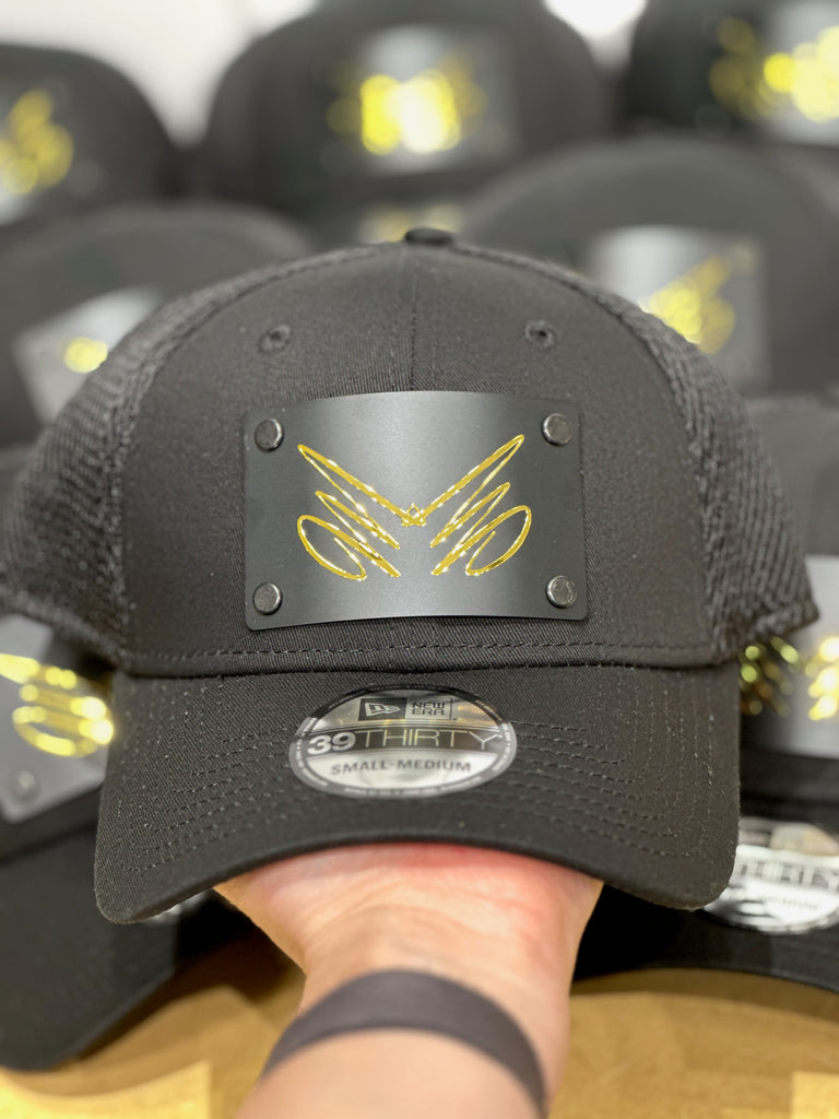 A custom black trucker hat of a metallic gold logo on a metal patch riveted on. Featuring the logo of Mirror, Mirror salon in Missoula, Montana.