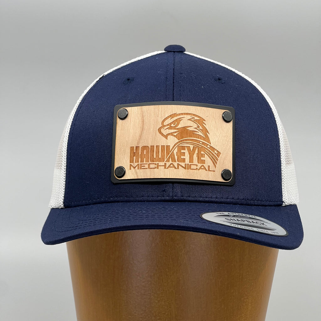 A Navy and white trucker hat with a wood plate patch of the logo for Hawkeye Mechanical