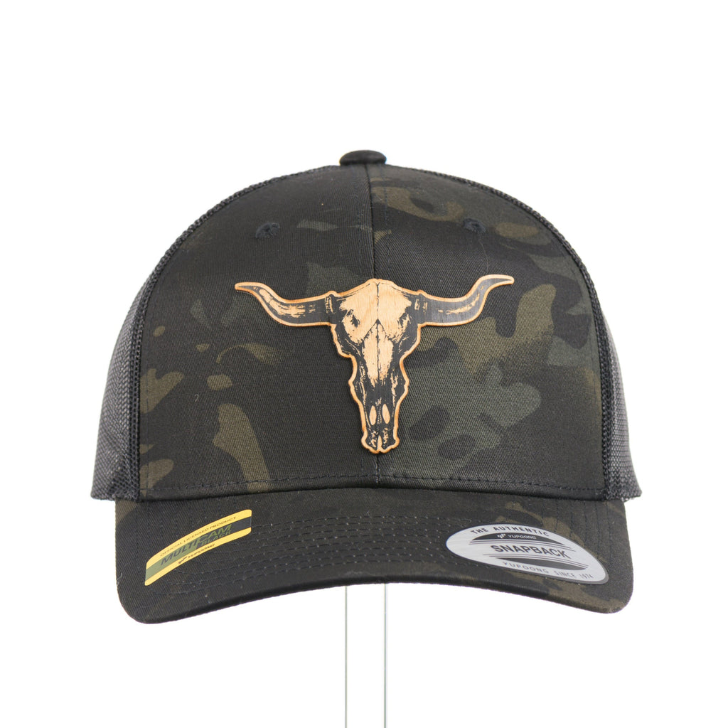 The front of a dark multicam trucker hat with a flexwood steer skull patch.
