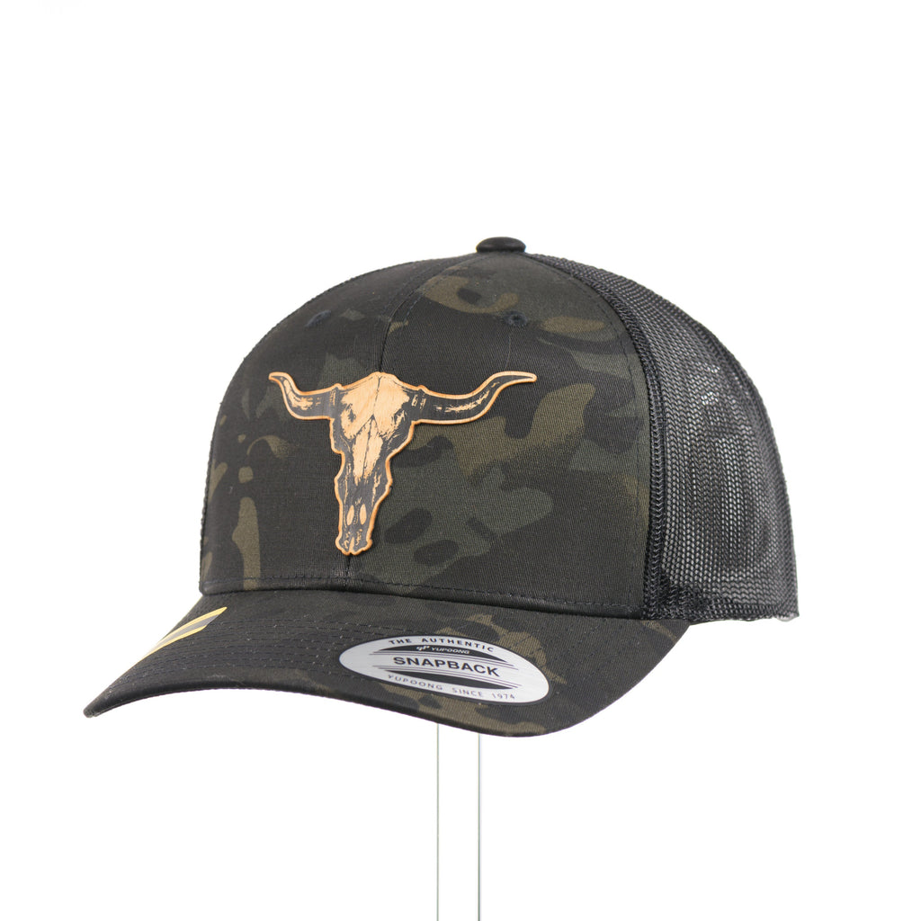 A dark multicam trucker hat with a flexwood steer skull patch at a quarter turn.