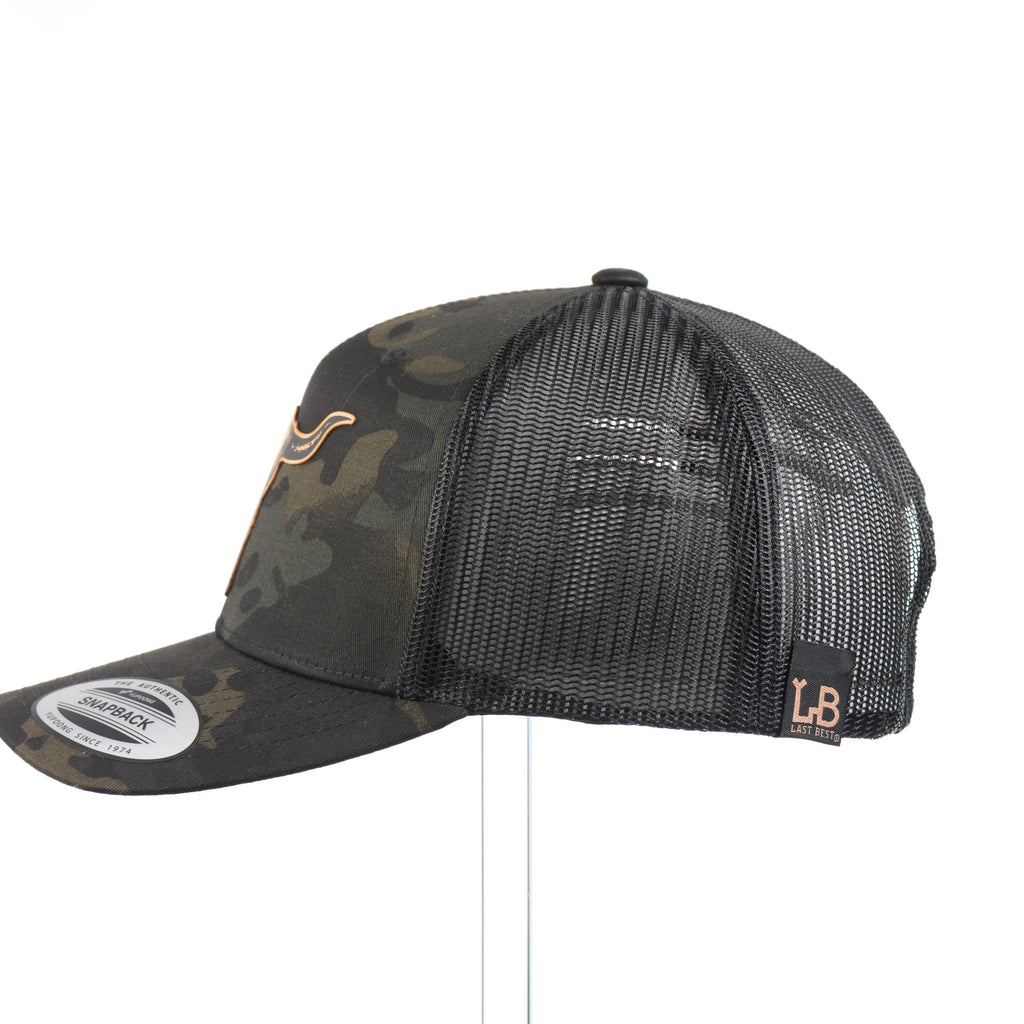 The side of a dark multicam trucker hat with a flexwood steer skull patch.