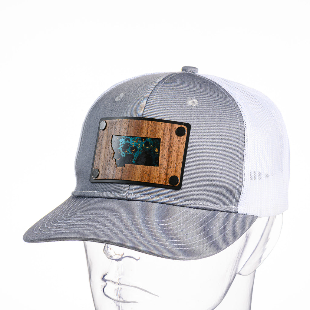 A Montana state walnut wood and teal copper plate patch riveted to a Heather grand white trucker cap
