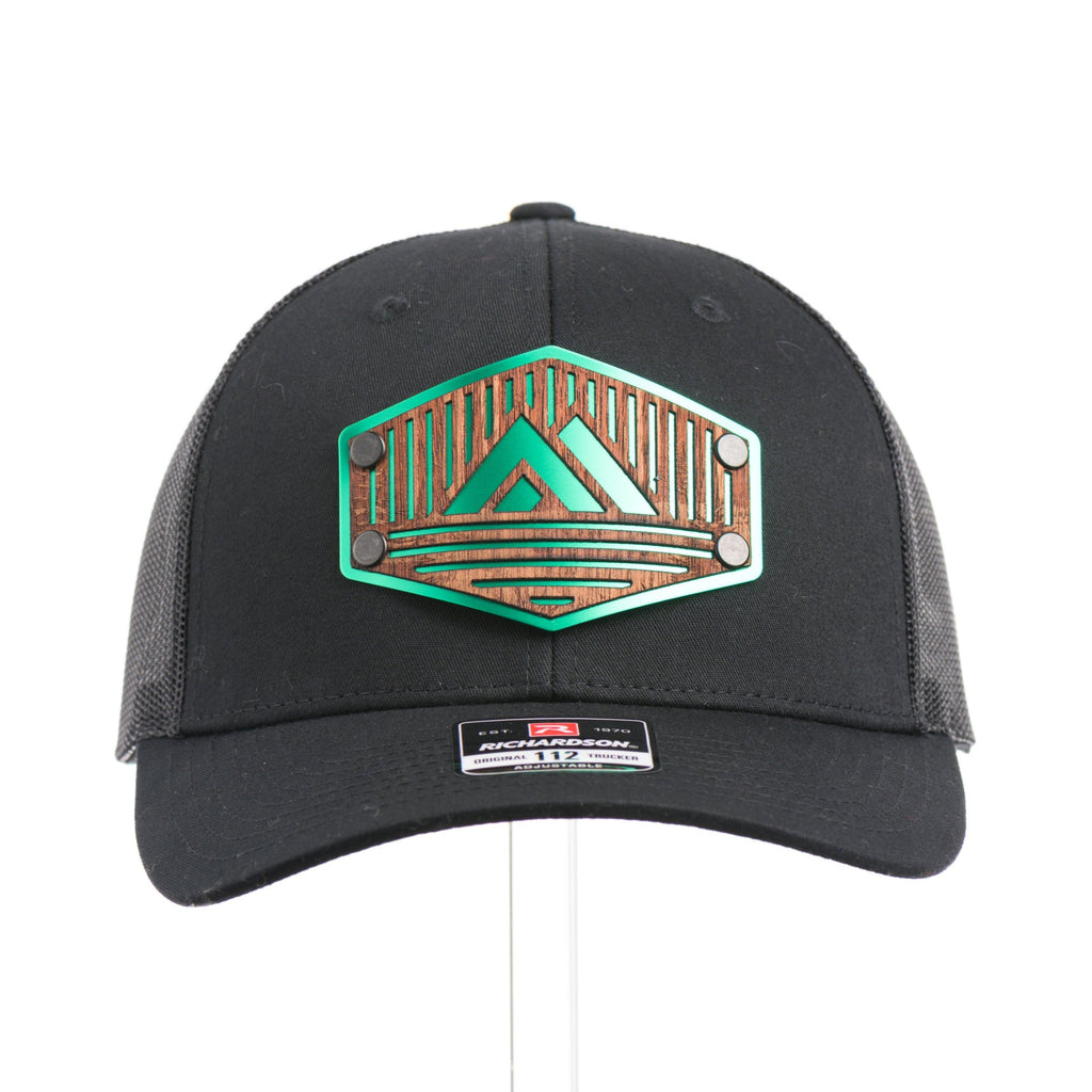 The front of a black Richardson trucker hat with an etched mahogany wood triangle camping patch on a green metal plate.