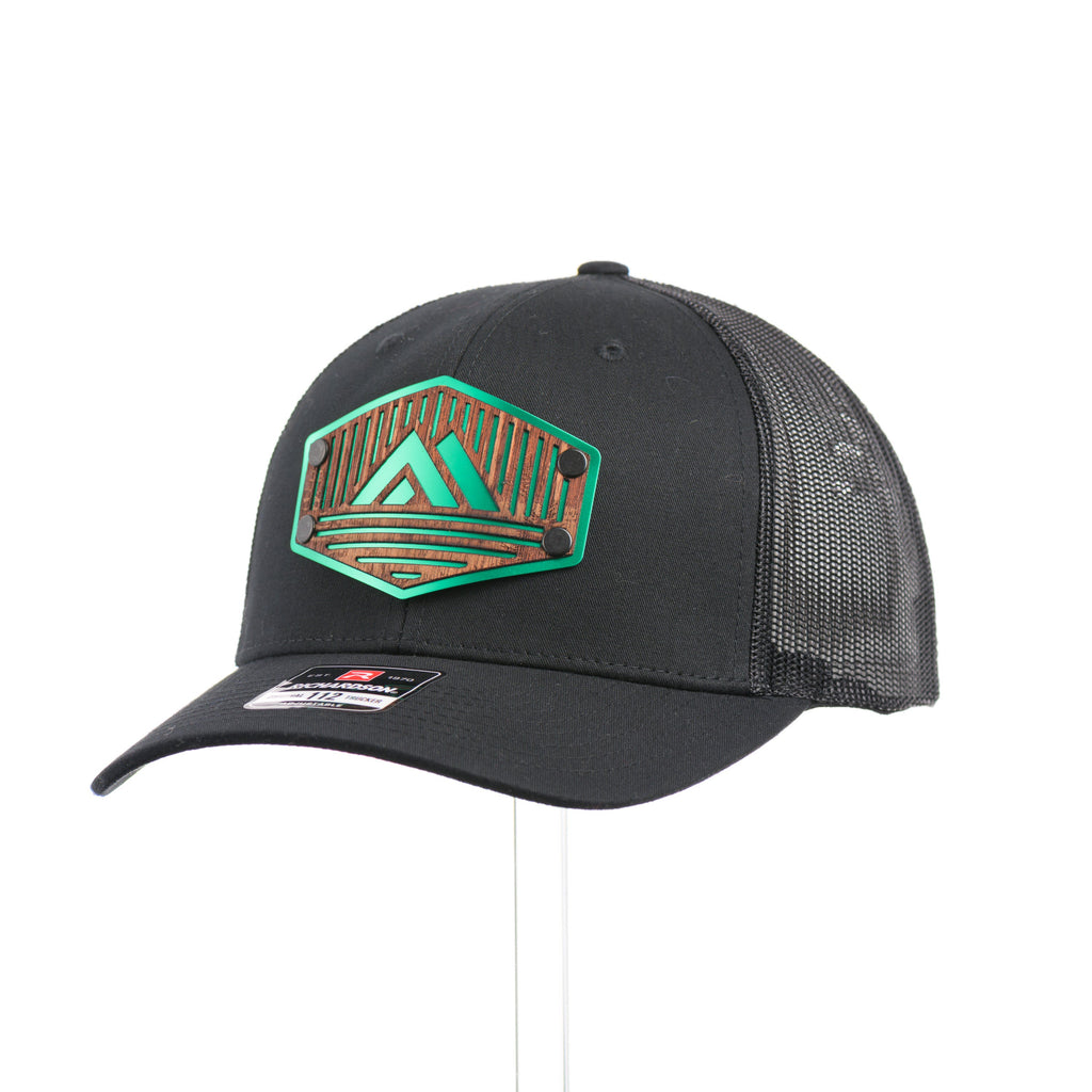 A black Richardson trucker hat with an etched mahogany wood triangle camping patch on a green metal plate at a quarter turn.