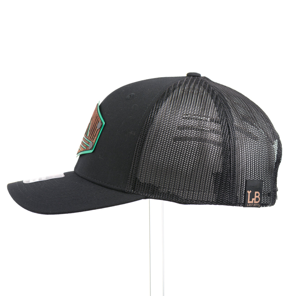 The side of a black Richardson trucker hat with an etched mahogany wood triangle camping patch on a green metal plate.