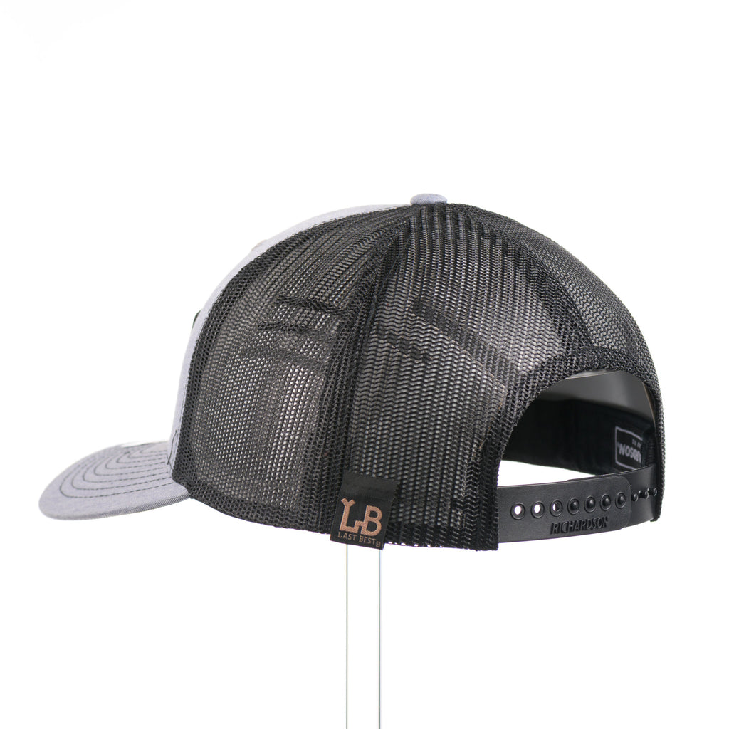 The back of a heather gray and black Richardson trucker hat with an etched walnut wood American flag and elk patch on a hexagon metal plate.