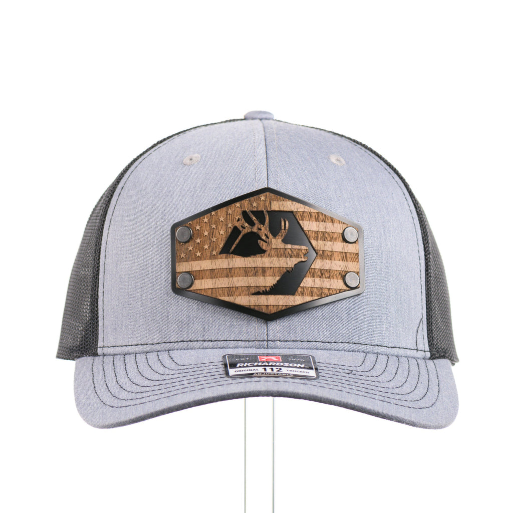 The front of a heather gray and black Richardson trucker hat with an etched walnut wood American flag and elk patch on a hexagon metal plate.