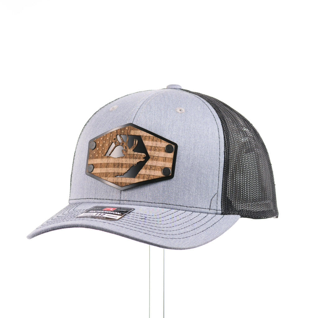 A heather gray and black Richardson trucker hat with an etched walnut wood American flag and elk patch on a hexagon metal plate at a quarter turn.