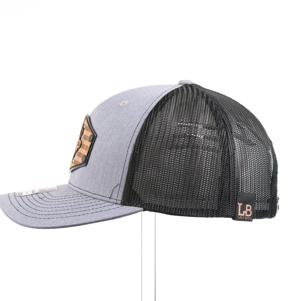 The side of a heather gray and black Richardson trucker hat with an etched walnut wood American flag and elk patch on a hexagon metal plate.