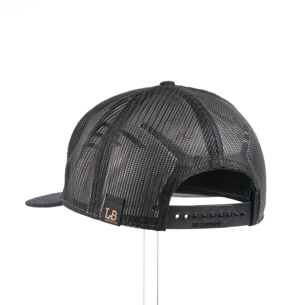 The back of a heather grey and black seven panel Richardson flat bill trucker hat with an evergreen trees leather patch thick stitched on.