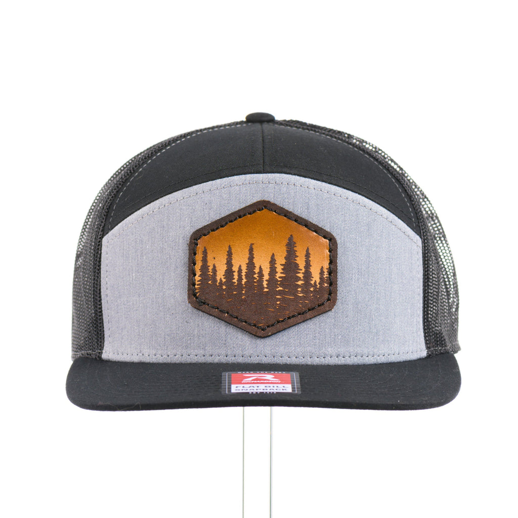 The front of a heather gray and black seven panel Richardson flat bill trucker hat with an evergreen trees leather patch thick stitched on.