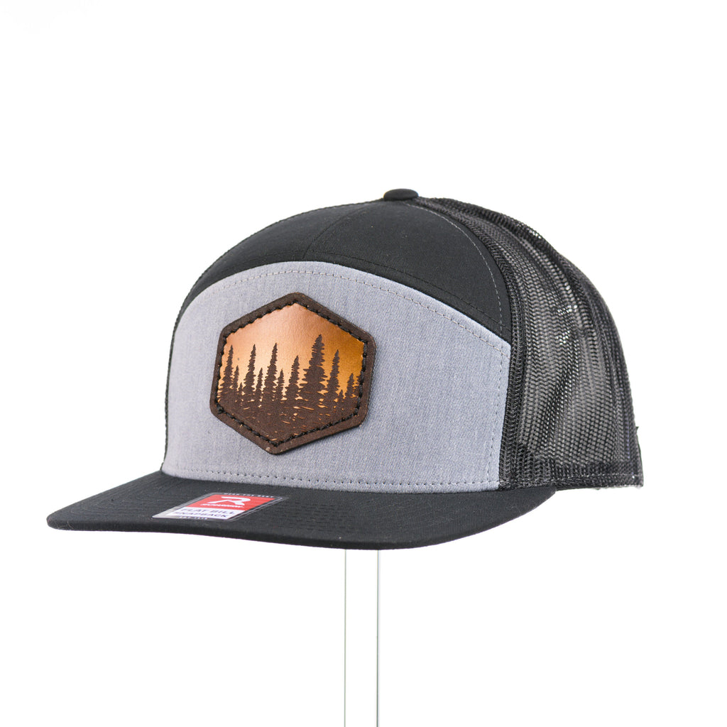 A heather grey and black seven panel Richardson flat bill trucker hat with an evergreen trees leather patch thick stitched on at a quarter turn.