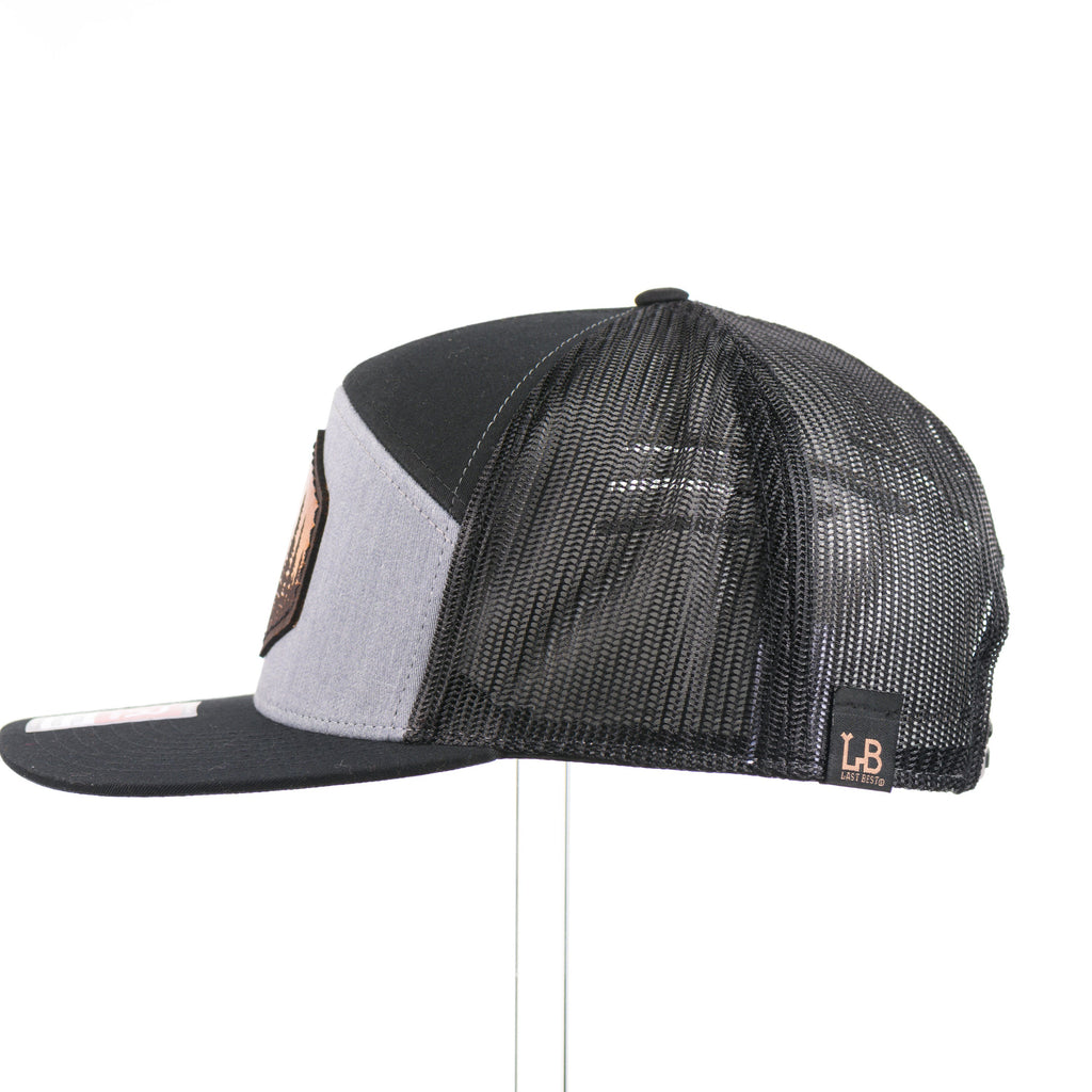 The side of a heather grey and black seven panel Richardson flat bill trucker hat with an evergreen trees leather patch thick stitched on.