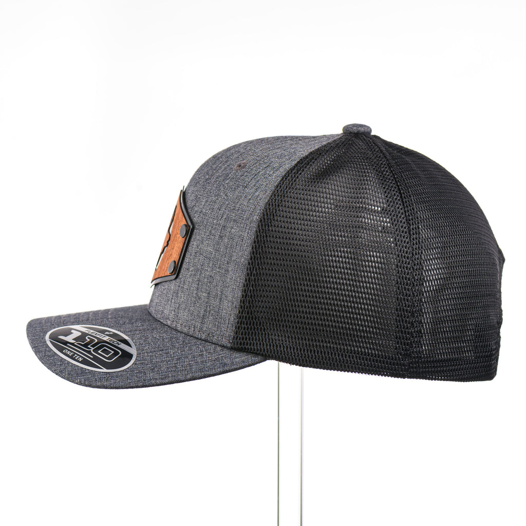 The side view of a dark heather gray and black mesh flexfit trucker hat with a mahogany wood elk cut out on a hexagon metal plate.