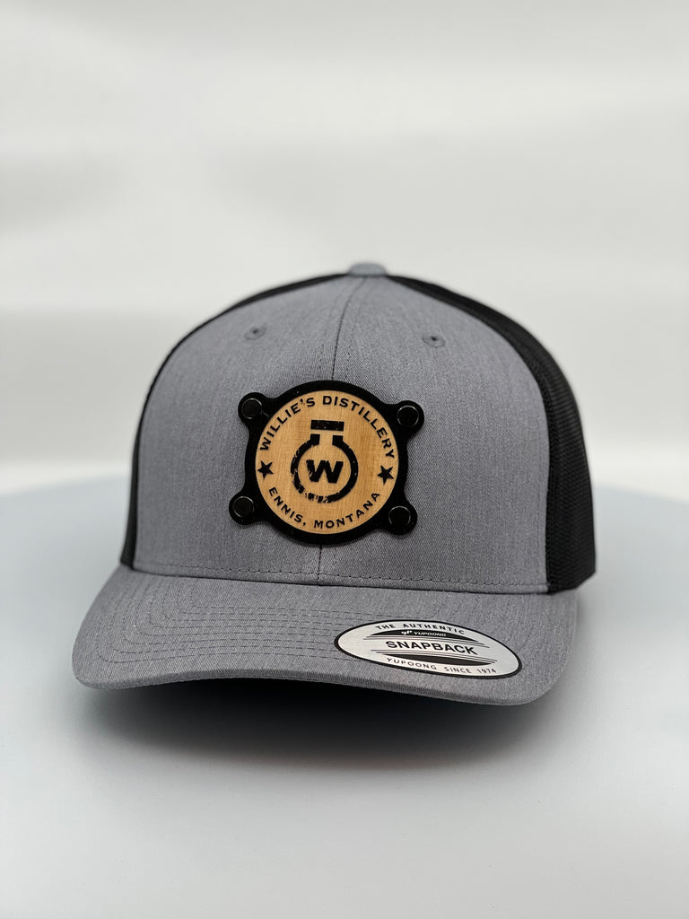 A custom heather grey and black trucker hat with a solid wood patch featuring the logo of Willie's Distillery
