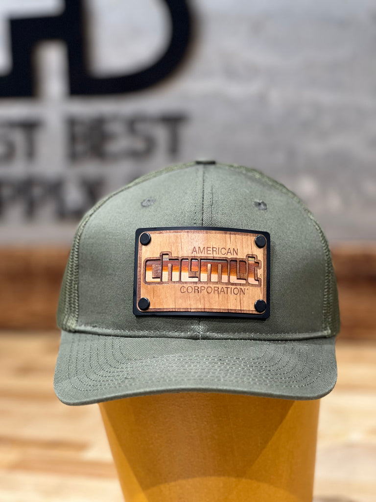 An olive green trucker hat with a wood patch made out of cherry wood veneer, copper and anodized aluminum featuring the logo of American Chemet Corporation