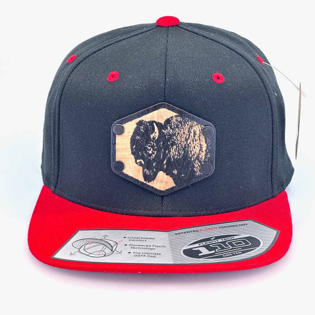 A solid wood patch of a bison head riveted to a black and red flex fit flat bill hat.
