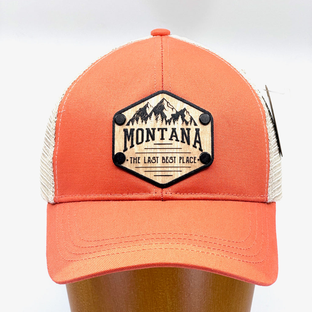 A solid wood patch riveted to a hat in the shape of a hexagon with the words, Montana, the last best place etched into it. On a peach and khaki trucker hat.