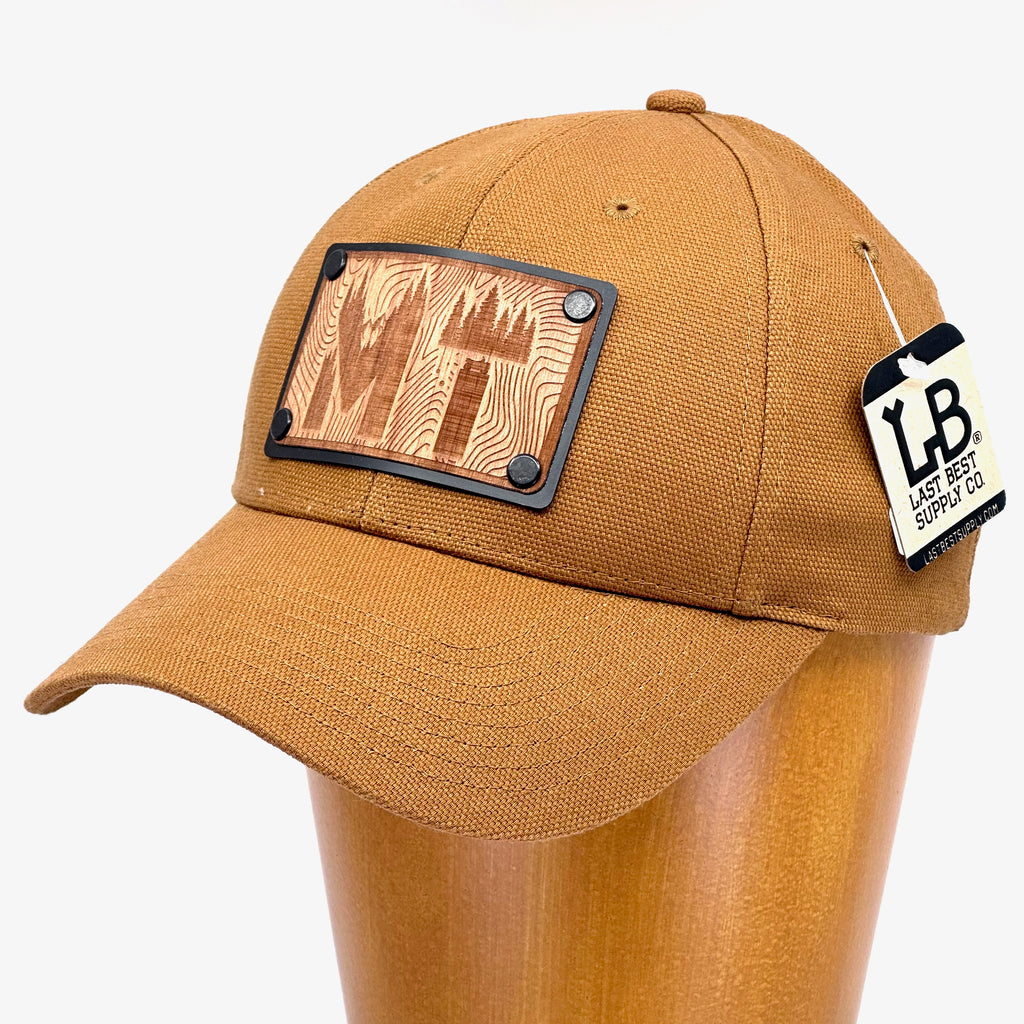 An etched MT, Montana wood plate patch riveted to a duck brown baseball hat.
