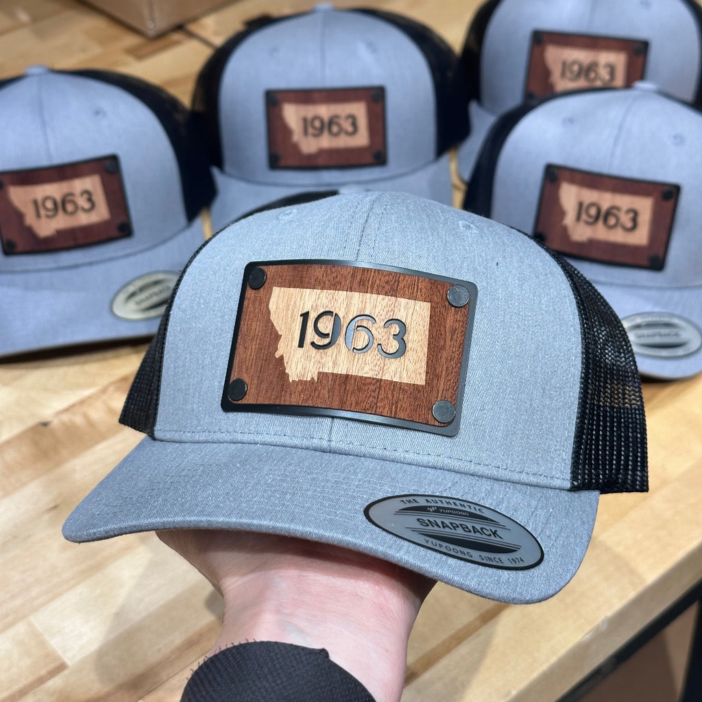 A heather grey and Black trucker hat with mahogany wood and aluminum patch featuring the state of Montana and 1963.