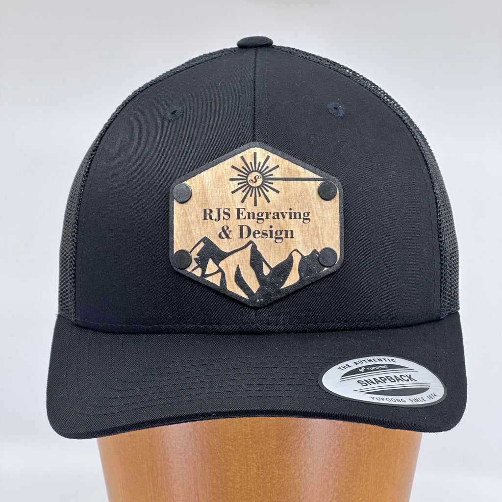 A custom black trucker hat with a solid wood patch featuring the logo of RJS Engraving & Design.