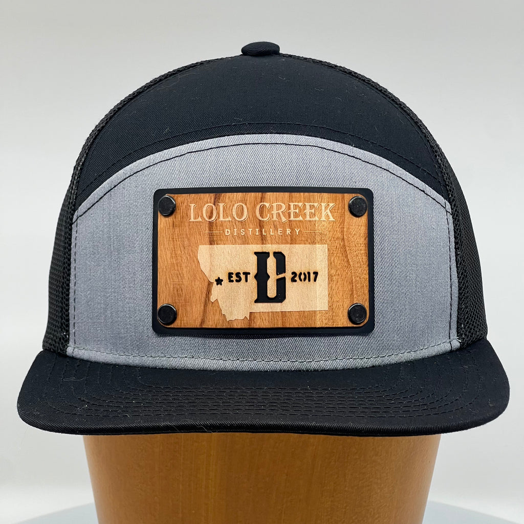 A custom flat bill trucker hat with a wood plate patch featuring the logo of Lolo Creek Distillery in Montana.