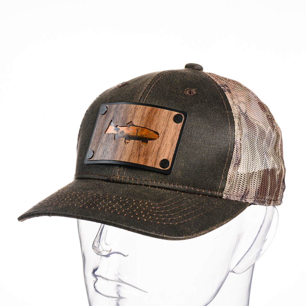 A trout silhouette cut out of walnut wood, with copper inlay on a plate patch riveted to a Kryptek trucker hat.