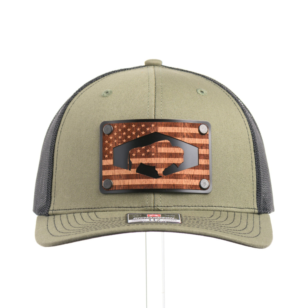The front of a loden green and black mesh Richardson trucker hat with an etched walnut wood American flag and bison cut out on a black metal patch.
