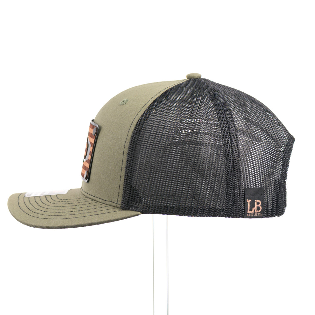 The side of a loden green and black mesh Richardson trucker hat with an etched  walnut wood American flag and bison cut out on a black metal patch.