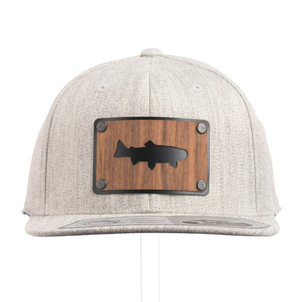 The front of a Heather Grey Flexfit Flat Bill hat with a cut out walnut wood trout patch on a black metal plate.