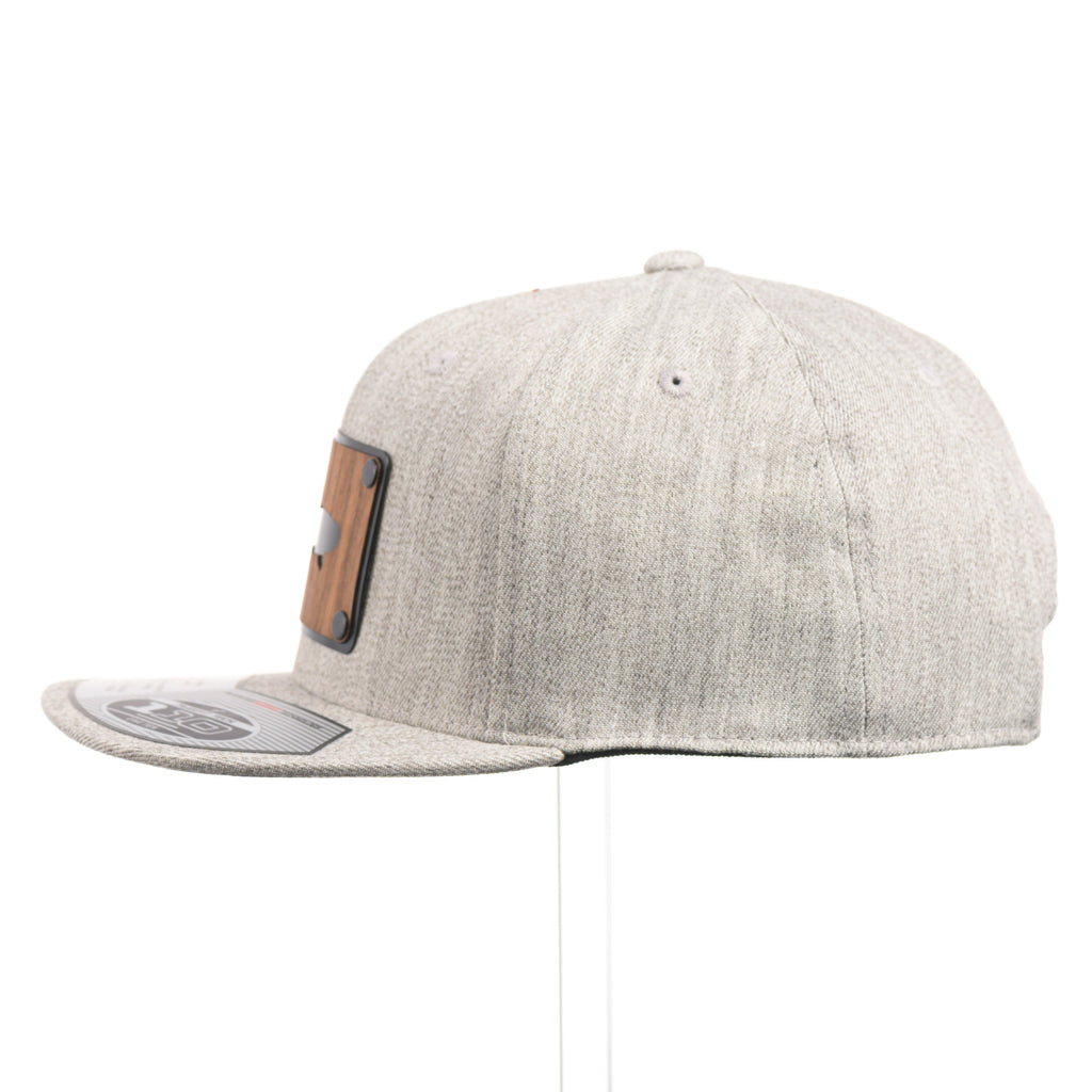 The side of a Heather Grey Flexfit Flat Bill hat with a cut out walnut wood trout patch on a black metal plate.