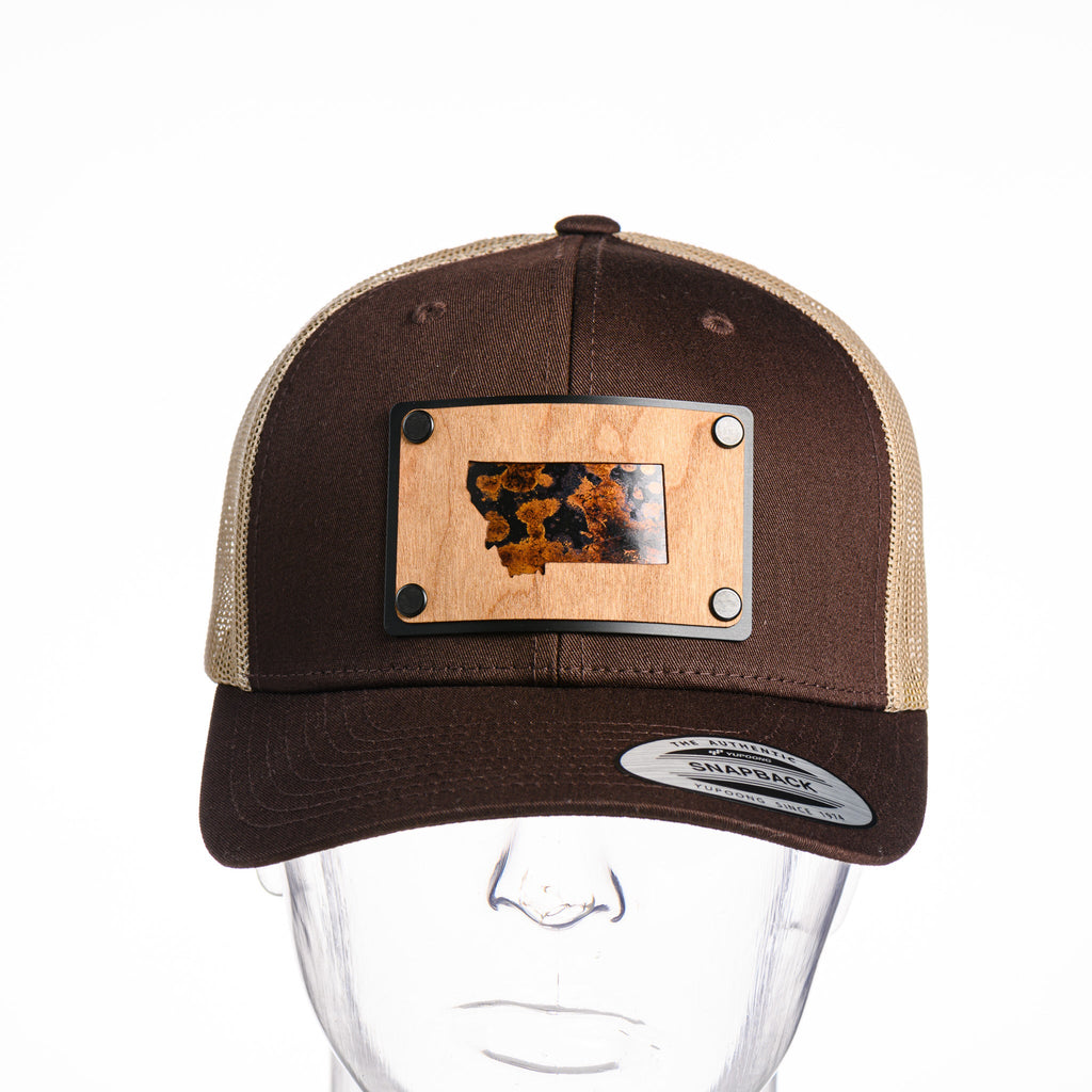 A Montana state Cherrywood and black and brown copper plate patch riveted to a chocolate brown and khaki trucker hat